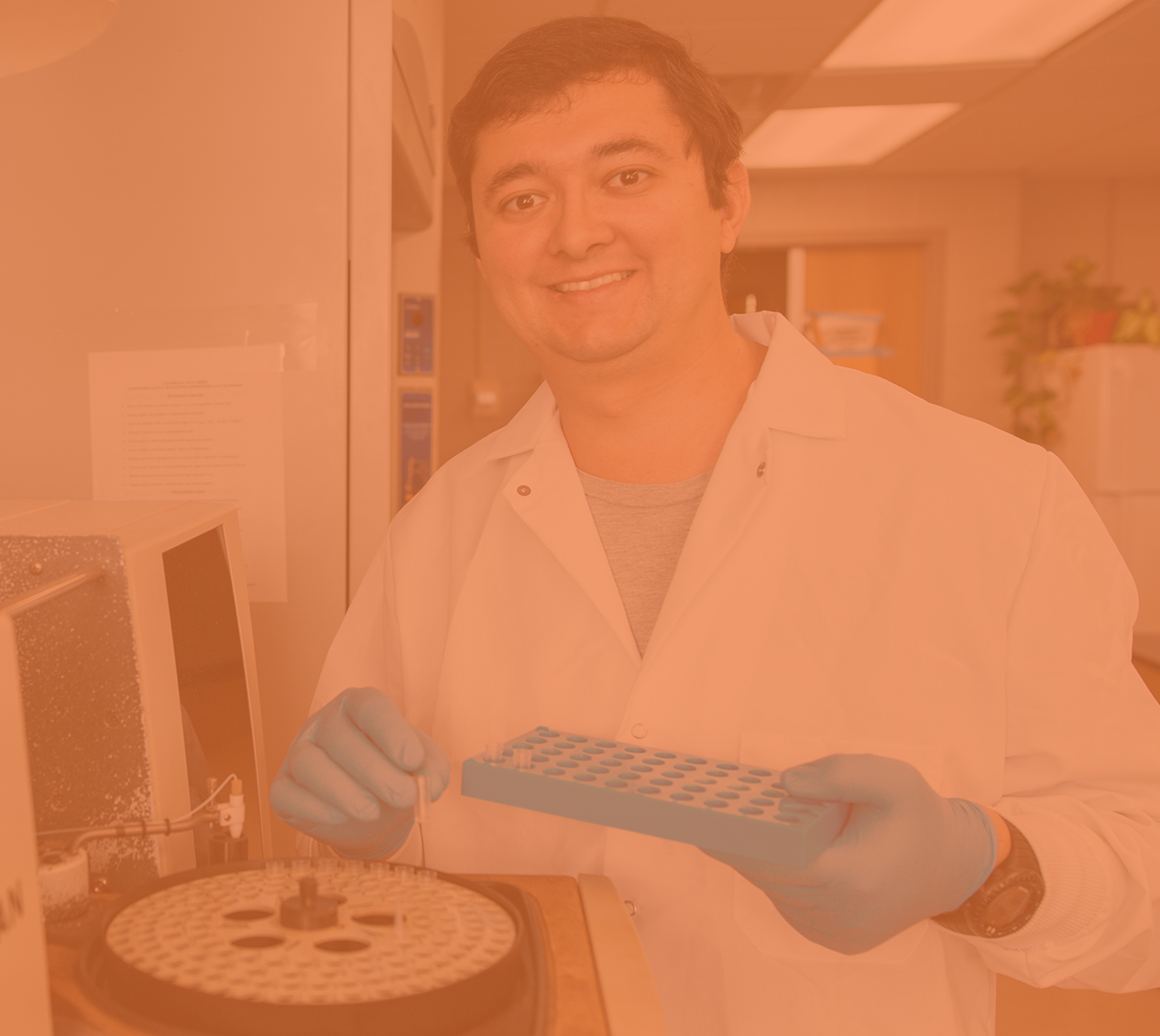 Male CALS student in a white lab coat and gloves smiles next to a centrifuge which he is loading. 