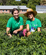 Two young women crouch in a field of green vegetation, wearing green shirts and smiling at the camera.