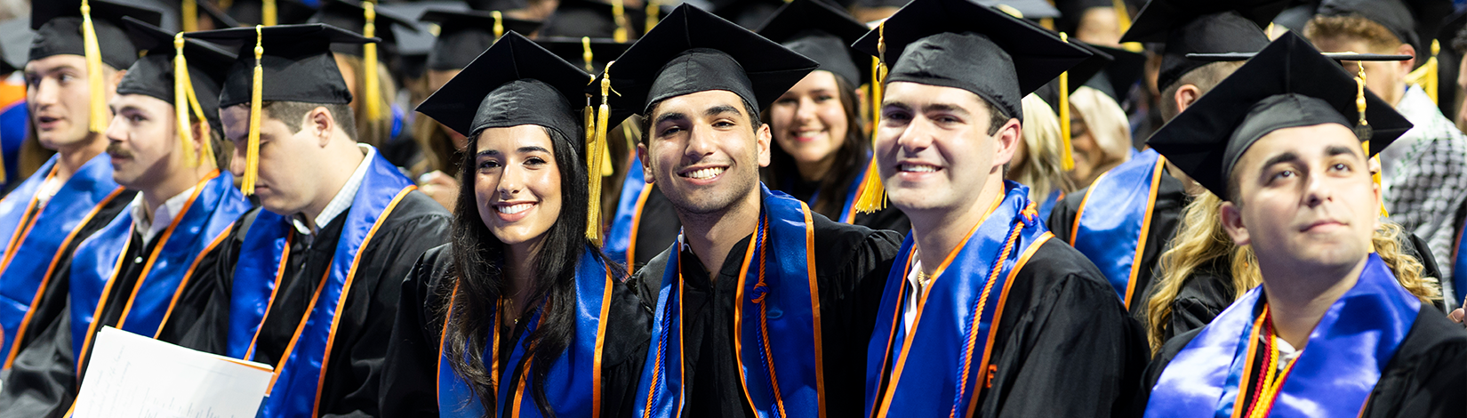 Several students sit in chairs at a graduation ceremony, wearing black robes and mortarboard caps with maize tassels.
