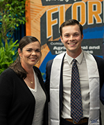 A student wearing a white graduation stole poses next to a woman wearing a white shirt and blazer.