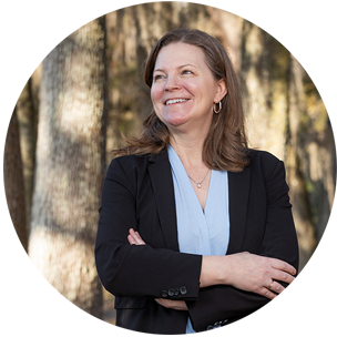 A woman in a black suit and light blue shirt standing outside, arms crossed, smiling and gazing over the viewer's