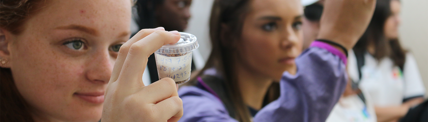 Student closely examines a plastic container with label. Students in the background are also examining containers.