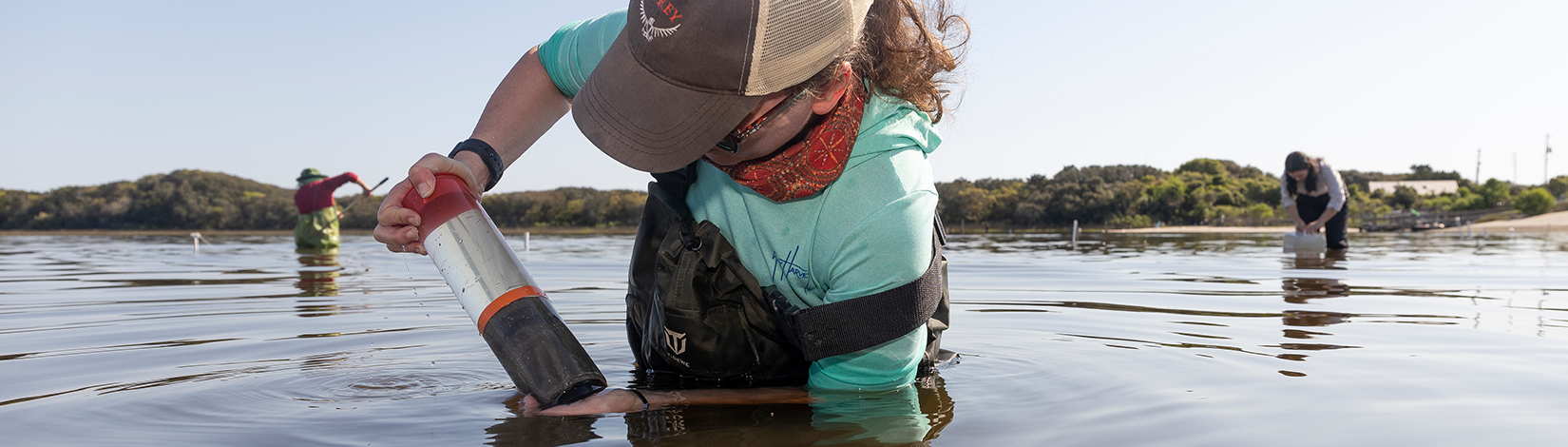 Student collects water sample while standing waist deep in water. In the background, two students are also examining water quality. 