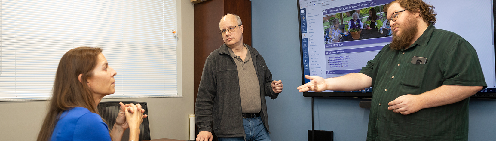 Two faculty members standing next to a screen give a presentation to a student sitting at a table.