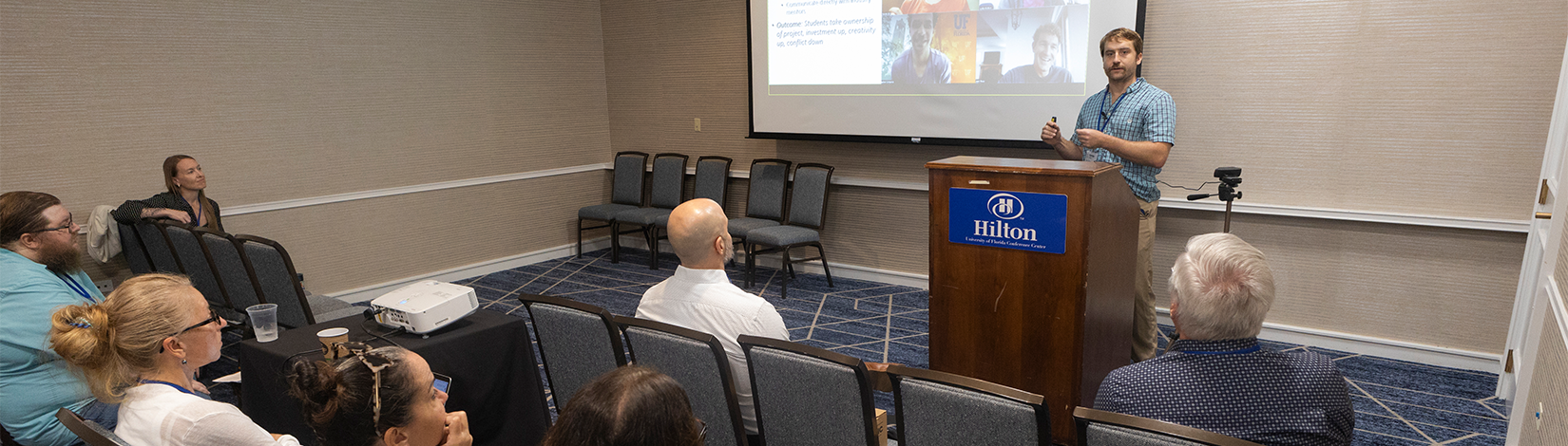 A man in a blue checked shirt stands at a podium, giving a presentation to a group seated in chairs.