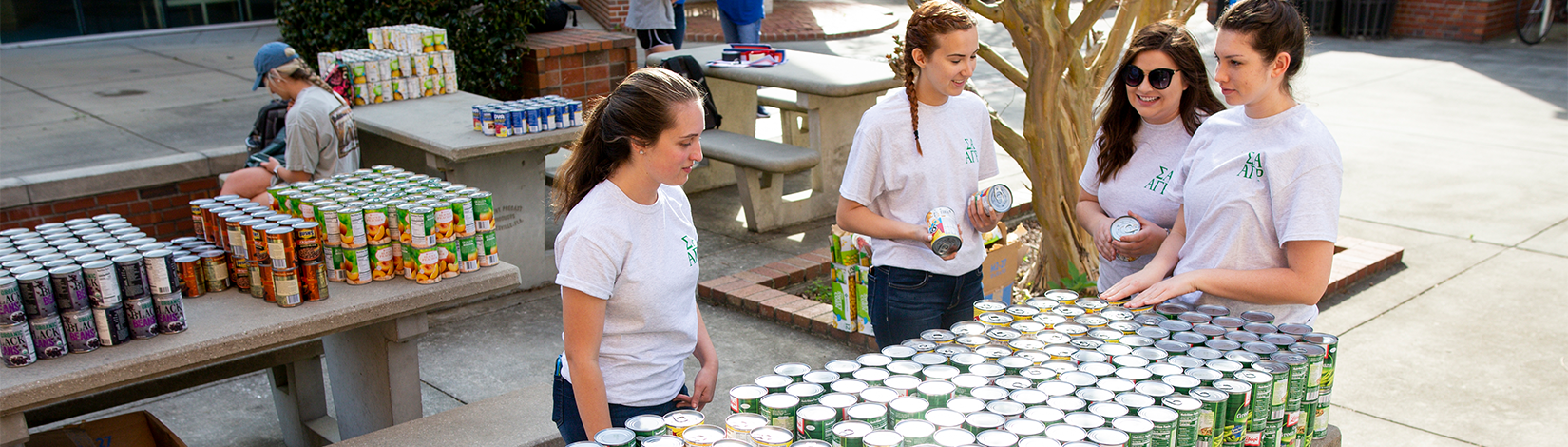Four female students gather around a table full of cans outside. 