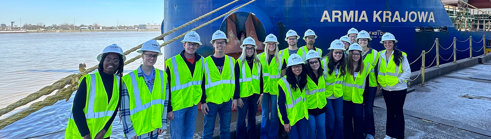 Students wearing neon yellow safety vests, hard hats, and safety glasses pose for a photo in front of a ship.