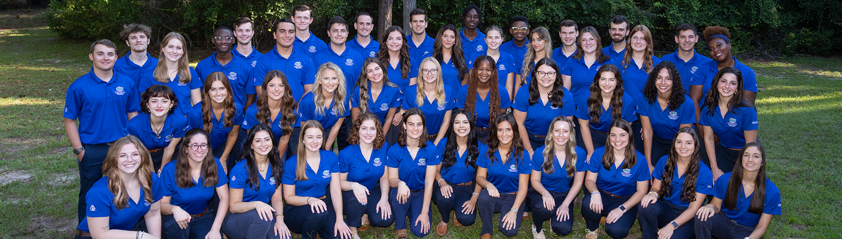 A large group of individuals wearing matching blue polo shirts outdoors on a grassy area with trees in the background.