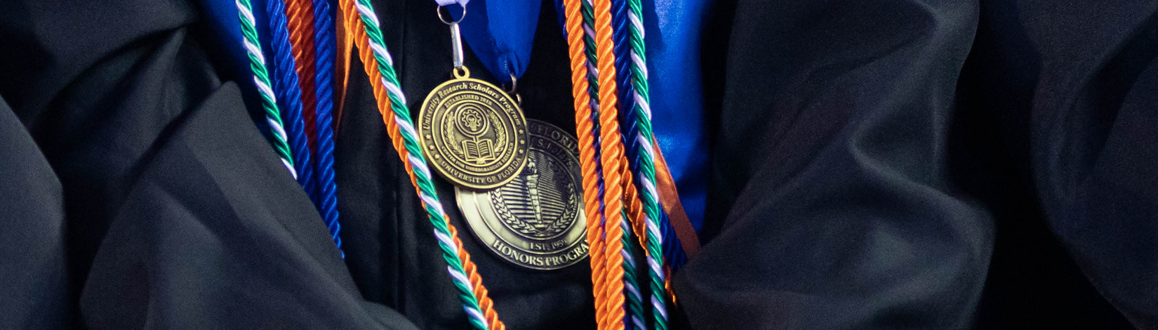 Cords and Medallions hang around graduating students robe. One Medallion reads Honors Program and the other reads University Research Scholars Program.  