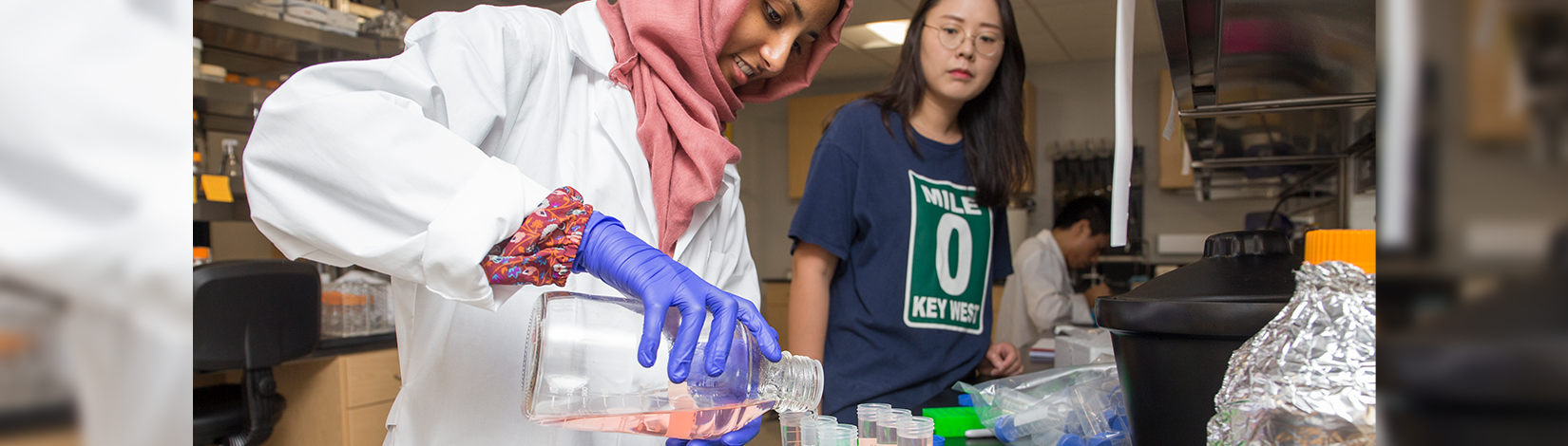 Female CALS student in white lab coat pours liquid into test tubes