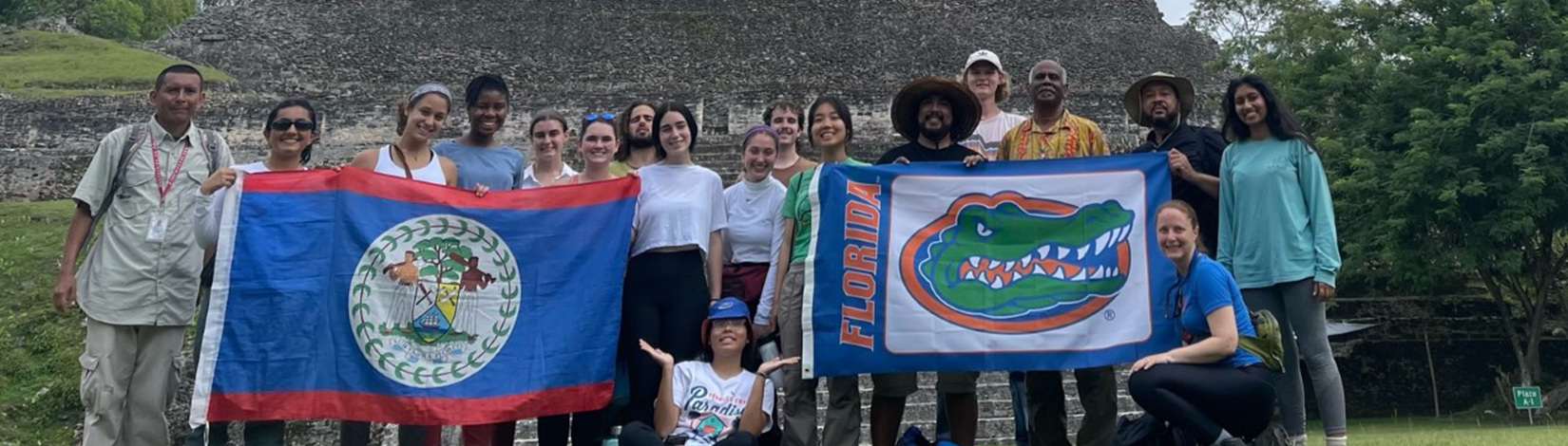 Students hold a Belize national flag on the left while students hold a Florida with a gator head flag on the right posing in front of an Aztec structure.