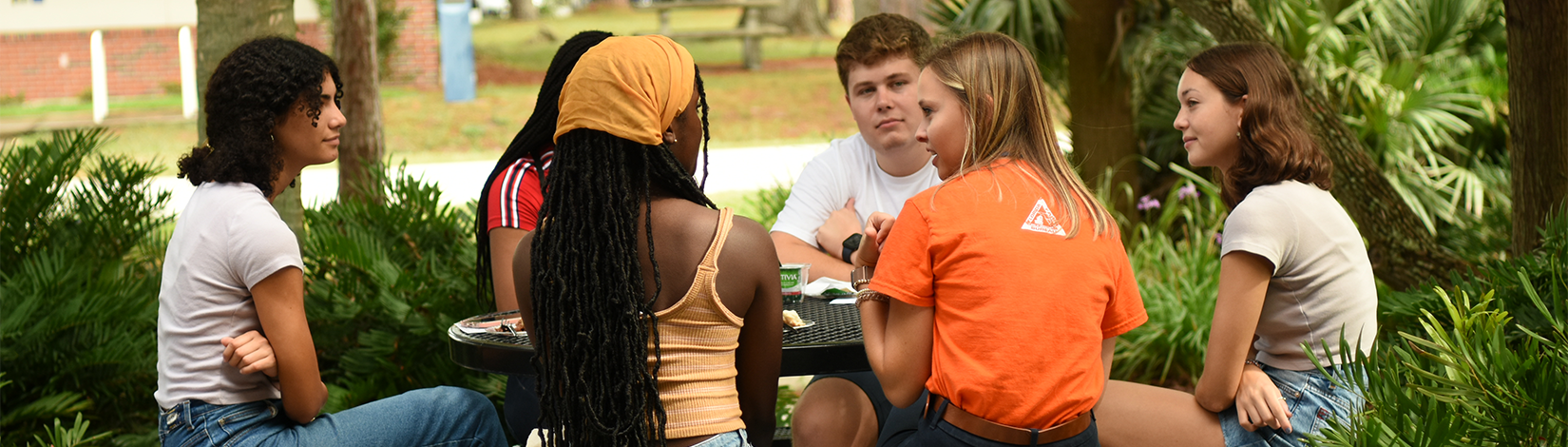 Six students sit and talk around a picnic table. 