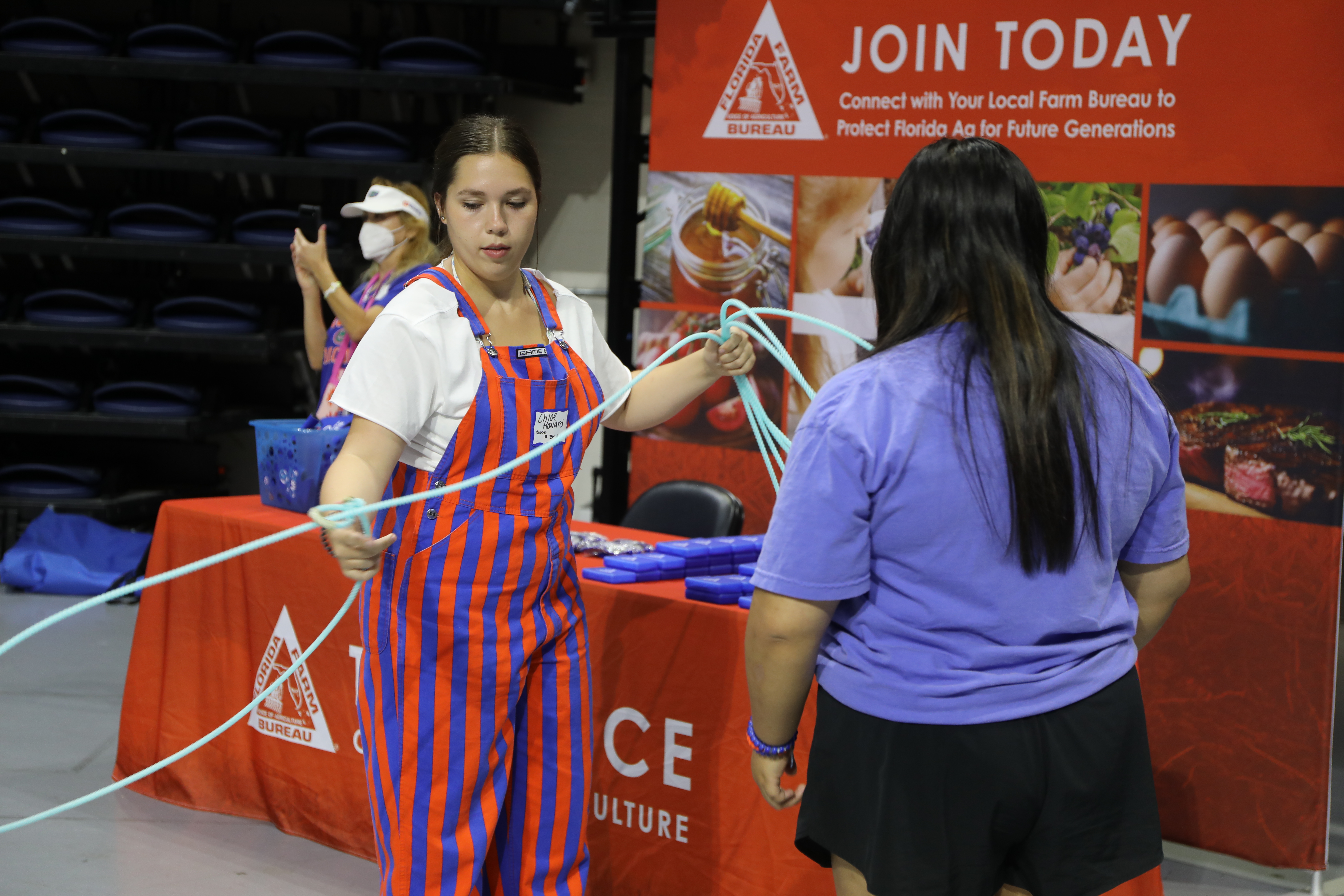 A student in orange and blue stripe overalls wraps up a lasso. Another student stands in front watching.
