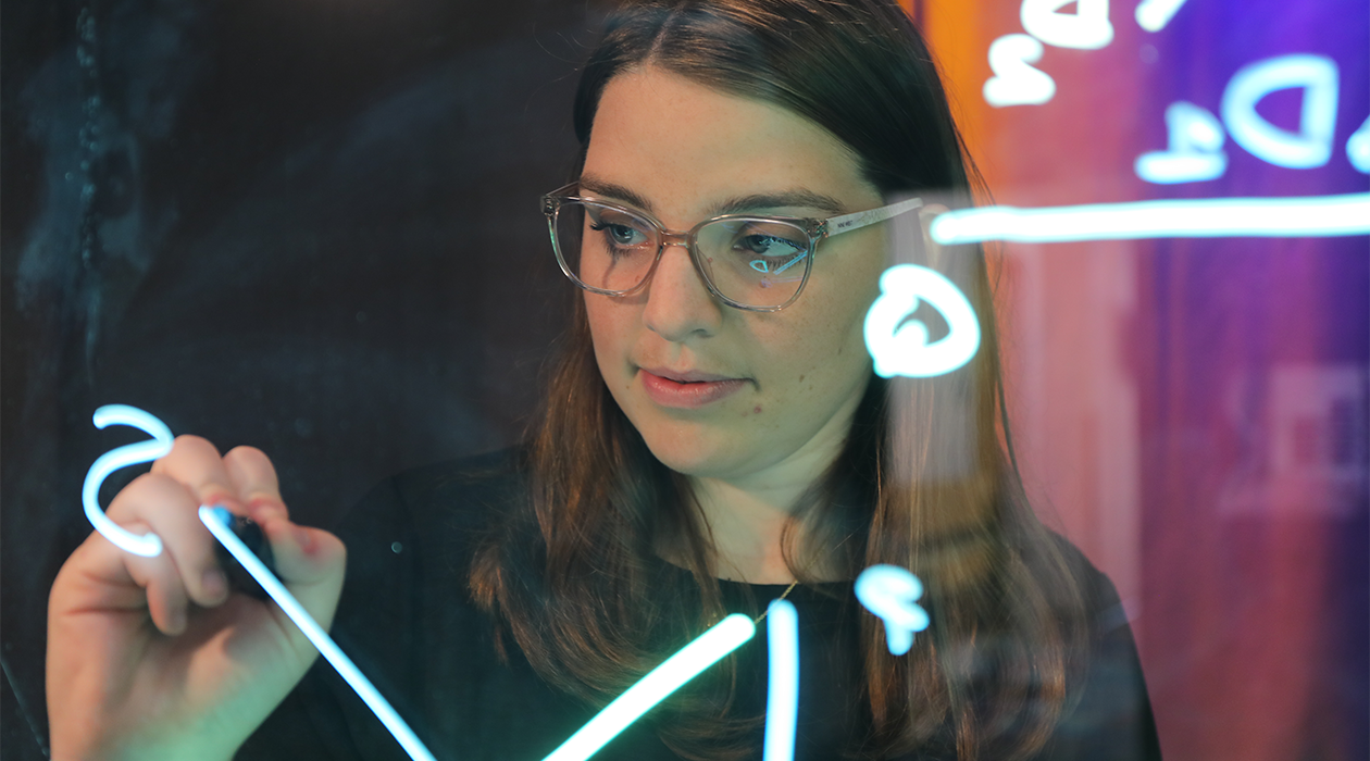 A student stands between the camera and a clear white board, writing a formula on the board.