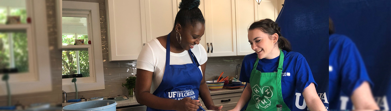 Two women in aprons collaborate in a kitchen, preparing food with various ingredients and utensils around them.